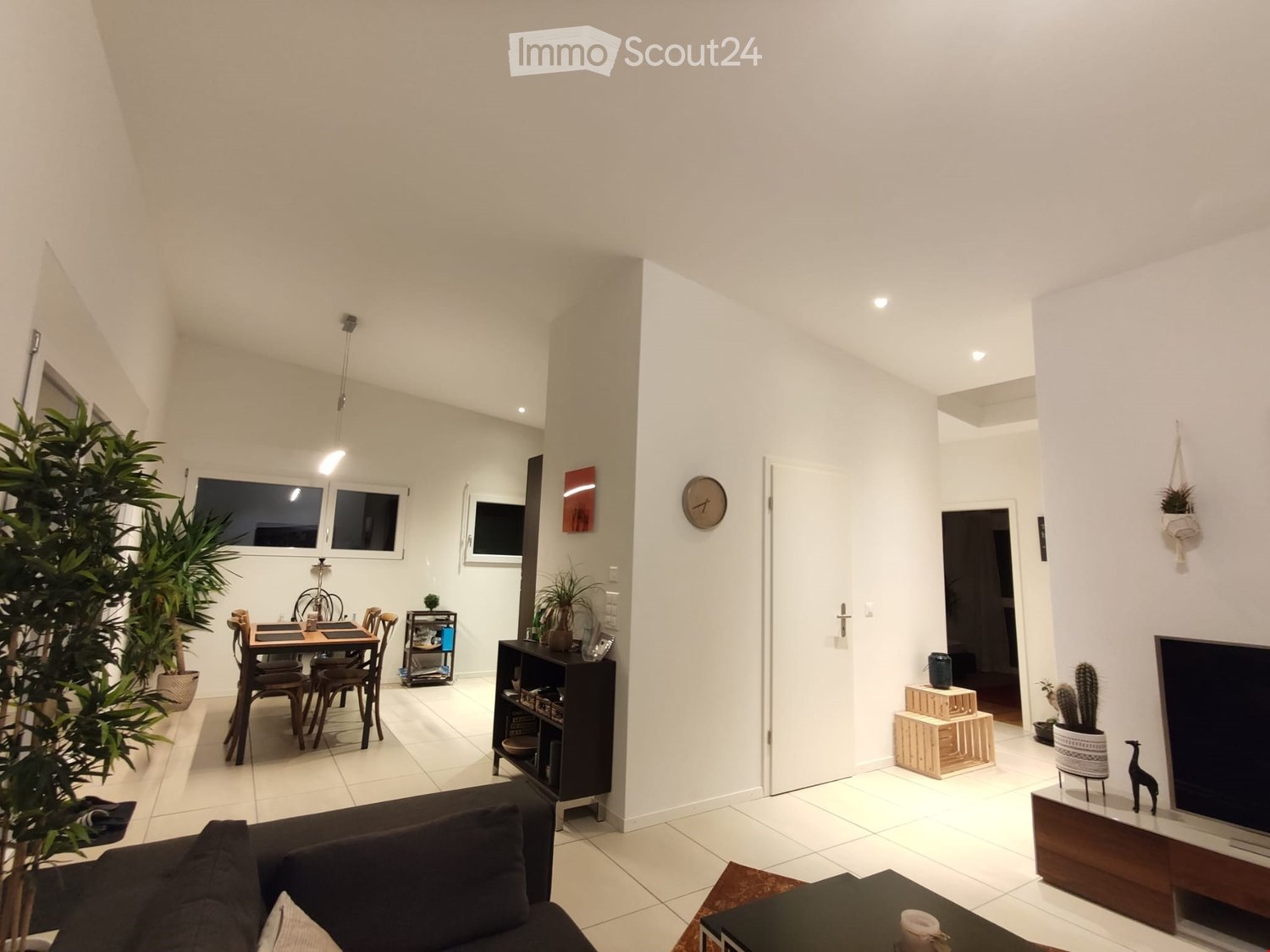 Modern living and dining area with white walls, tiled floor, and large windows. Features a dark-colored sofa, coffee table, dining table with chairs, shelves, and decorative plants.