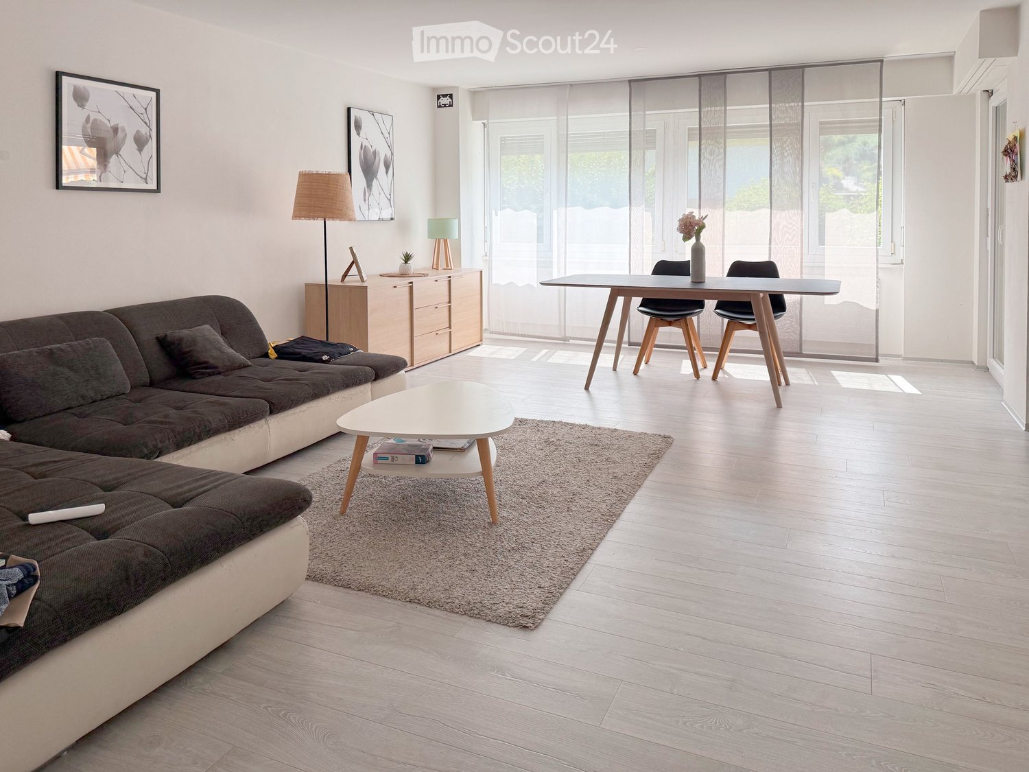 Modern living room with light wood flooring, large windows with blinds, a grey sectional sofa, a white coffee table, a dining table with chairs, and decorative items including a floor lamp and picture frames.