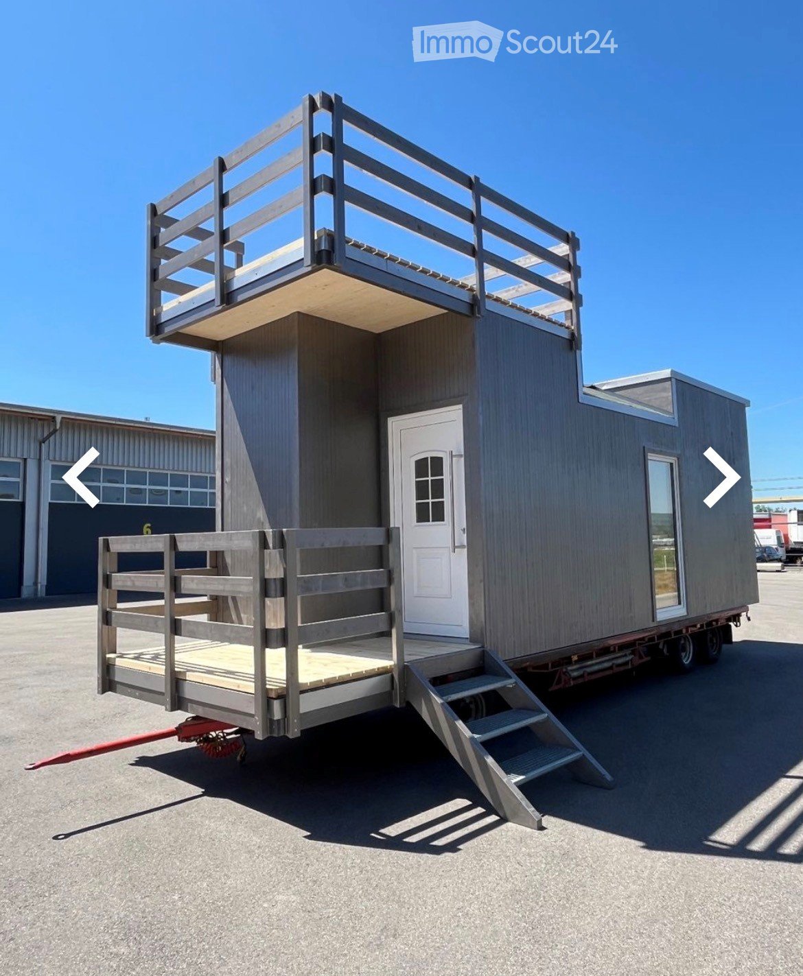 A modern, two-story tiny home on wheels with a wooden deck and staircase, featuring a white door and large windows.
