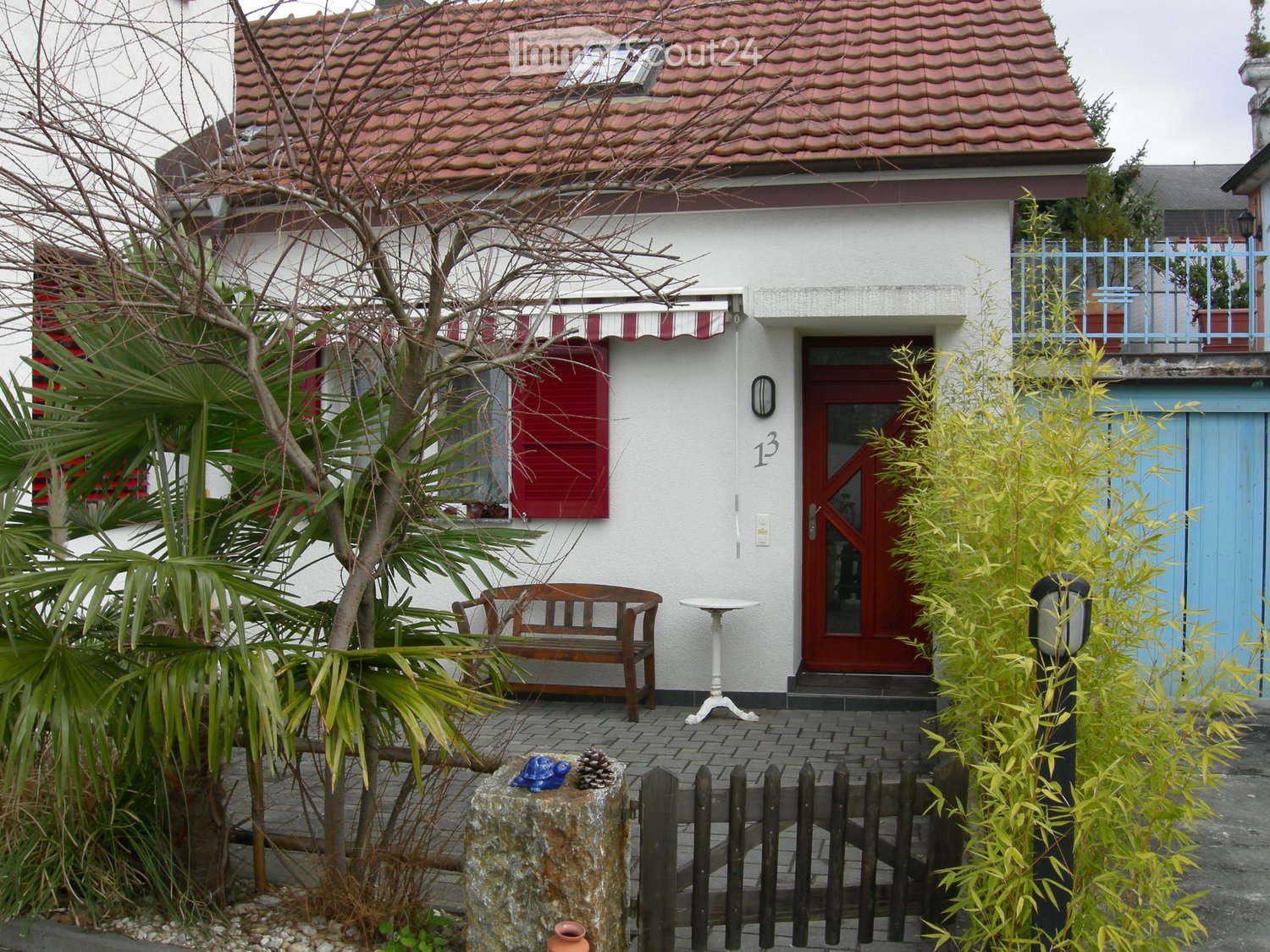 Two-story house with red-tiled roof, white walls, red shutters, and a small balcony with a railing. The entrance features a red door with a glass panel, and the house number "13" is displayed on the wall. The surrounding area includes a small garden with