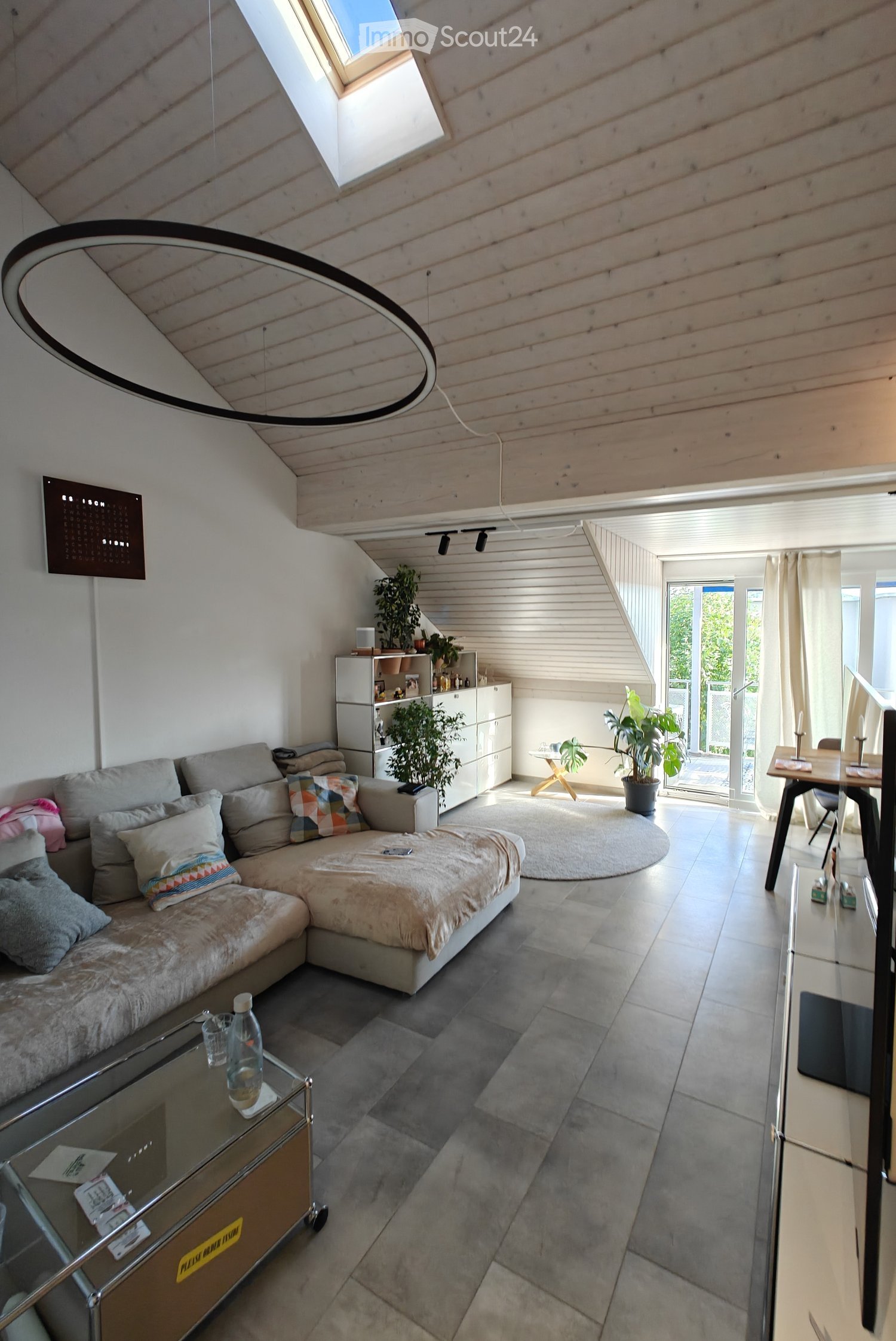 Modern living room with a grey sectional sofa, coffee table, dining table, chairs, and a sleek cabinet. The room features a skylight and large windows, with a round rug and decorative pillows on the sofa.