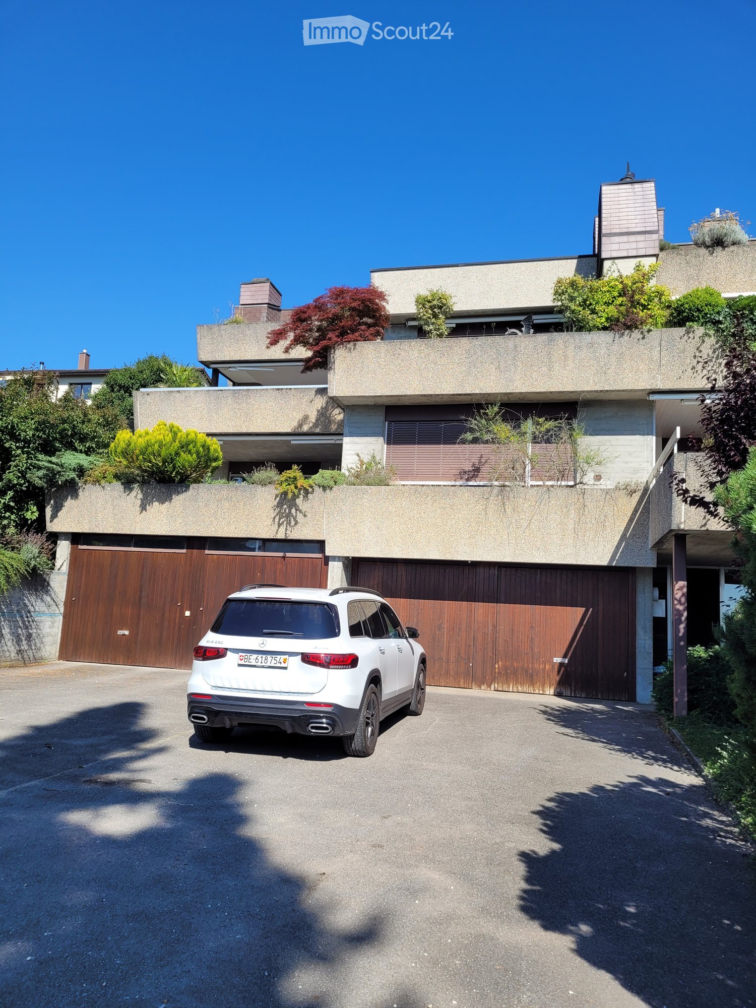 Modern two-story house with a flat roof, large wooden garage doors, and a white SUV parked in front. The house features a raised ground floor and is surrounded by greenery and plants.