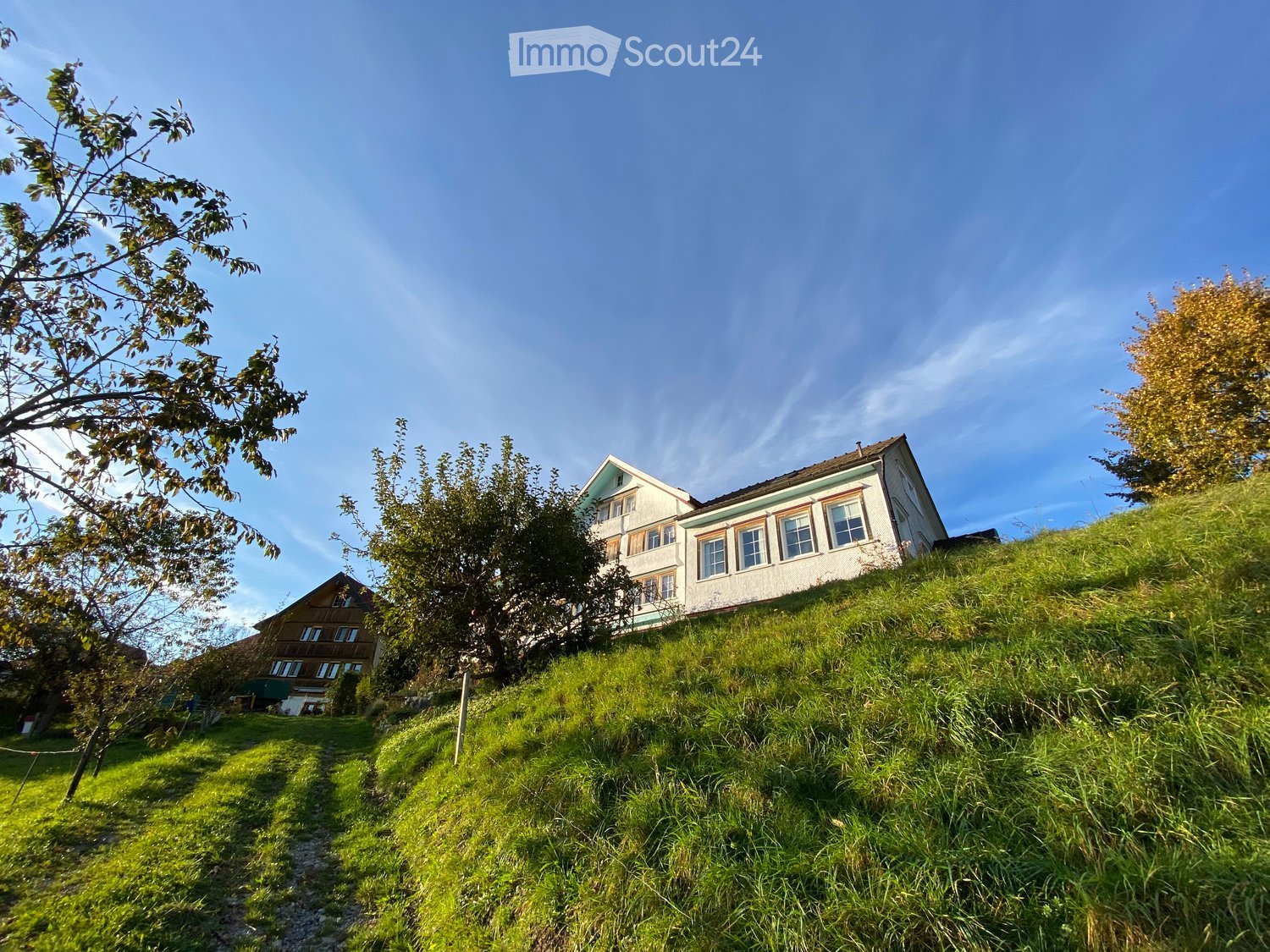 Two-story house with white walls and multiple windows, situated on a grassy hill with trees and a clear blue sky in the background.