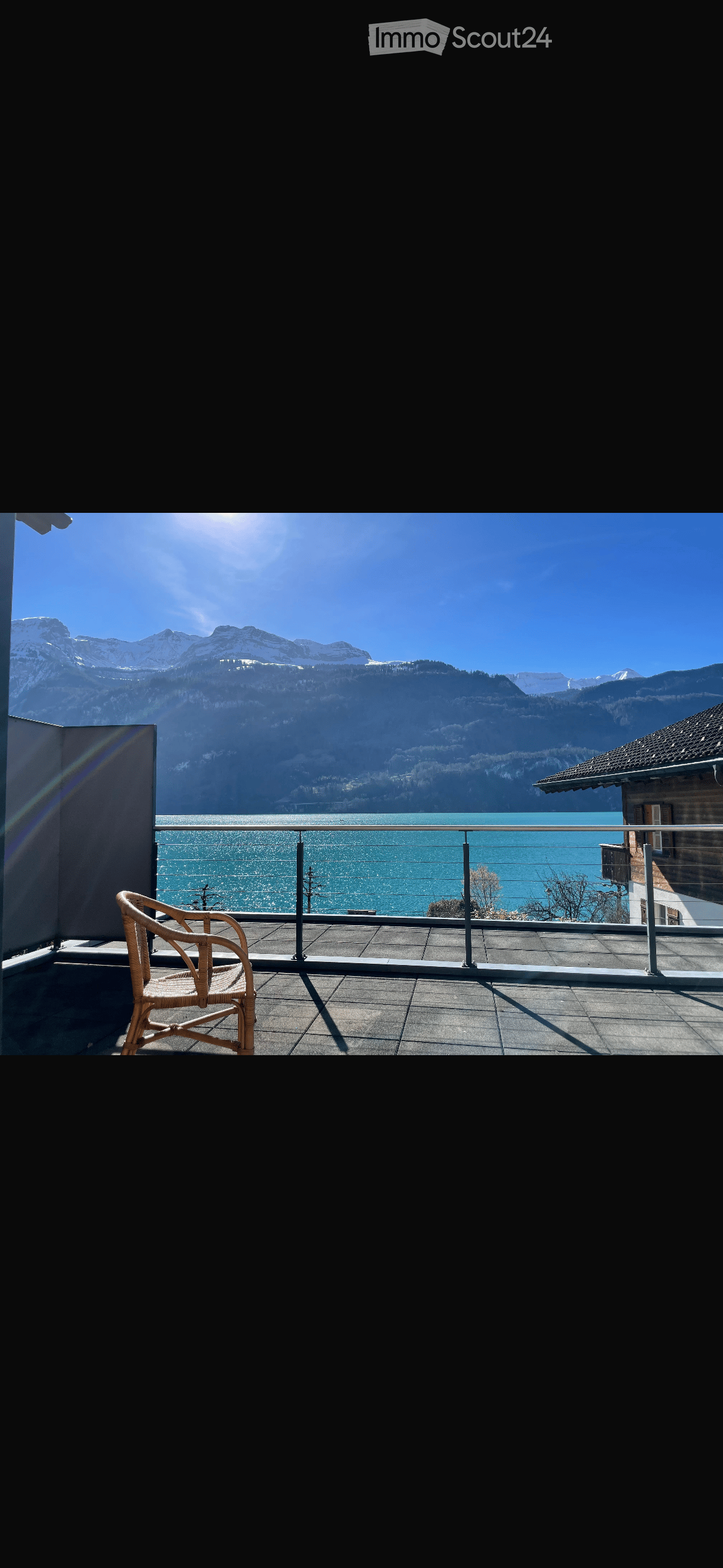 Balcony with a view of a lake and mountains, featuring a single wicker chair and a metal railing.