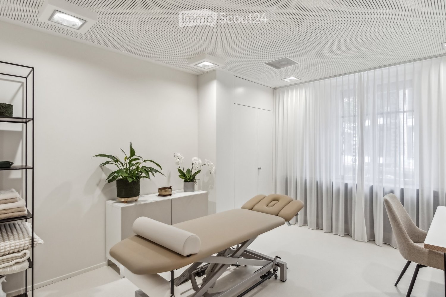 Modern, minimalist room with a massage table, a small table with a plant and decorative items, a chair, and a shelf with towels.