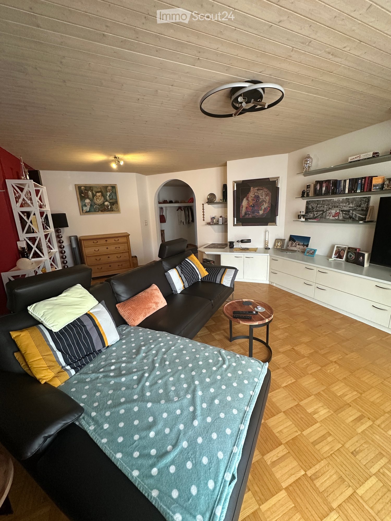 Modern living room with a black leather sofa, colorful pillows, a coffee table, a bookshelf, and a cabinet. The room features a blonde wood floor and a large window with a view.