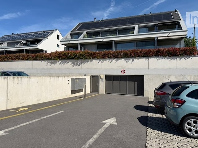 Modern multi-story building with solar panels on the roof, garage entrance, and adjacent parking area with several cars.