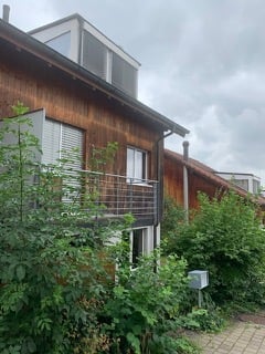 Two-story wooden building with a flat roof and balcony, surrounded by greenery.