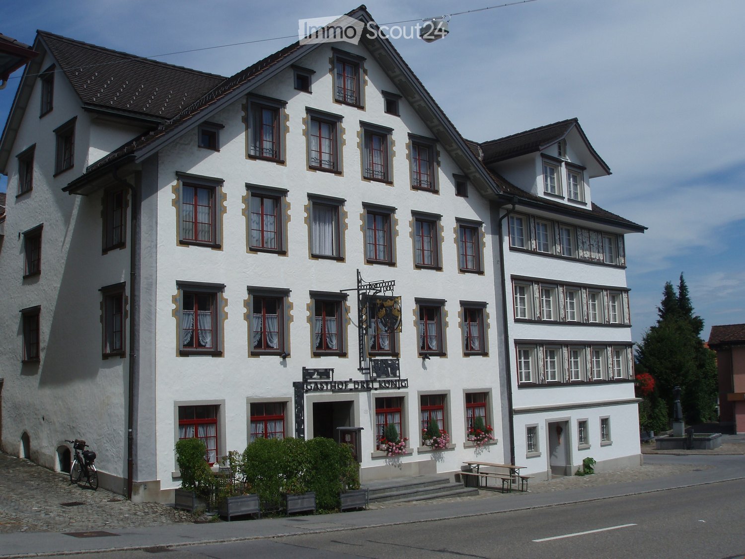 A three-story white building with red-framed windows and decorative trim, featuring a sign reading "Gasthof zur Krone" and a small outdoor seating area with tables and benches.