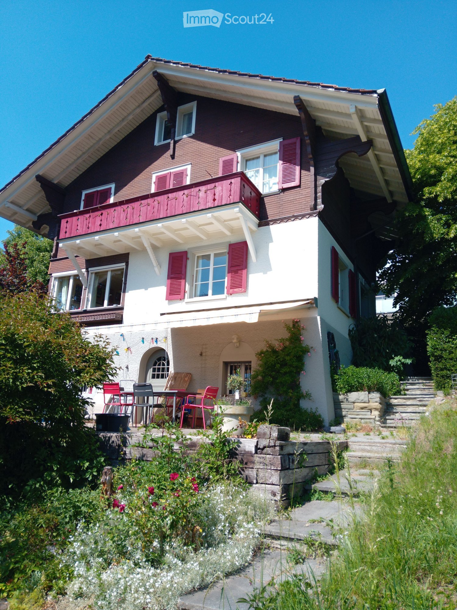 Two-story house with red brick and white siding, multiple windows with shutters, a balcony on the upper level, and a small patio area with outdoor furniture.