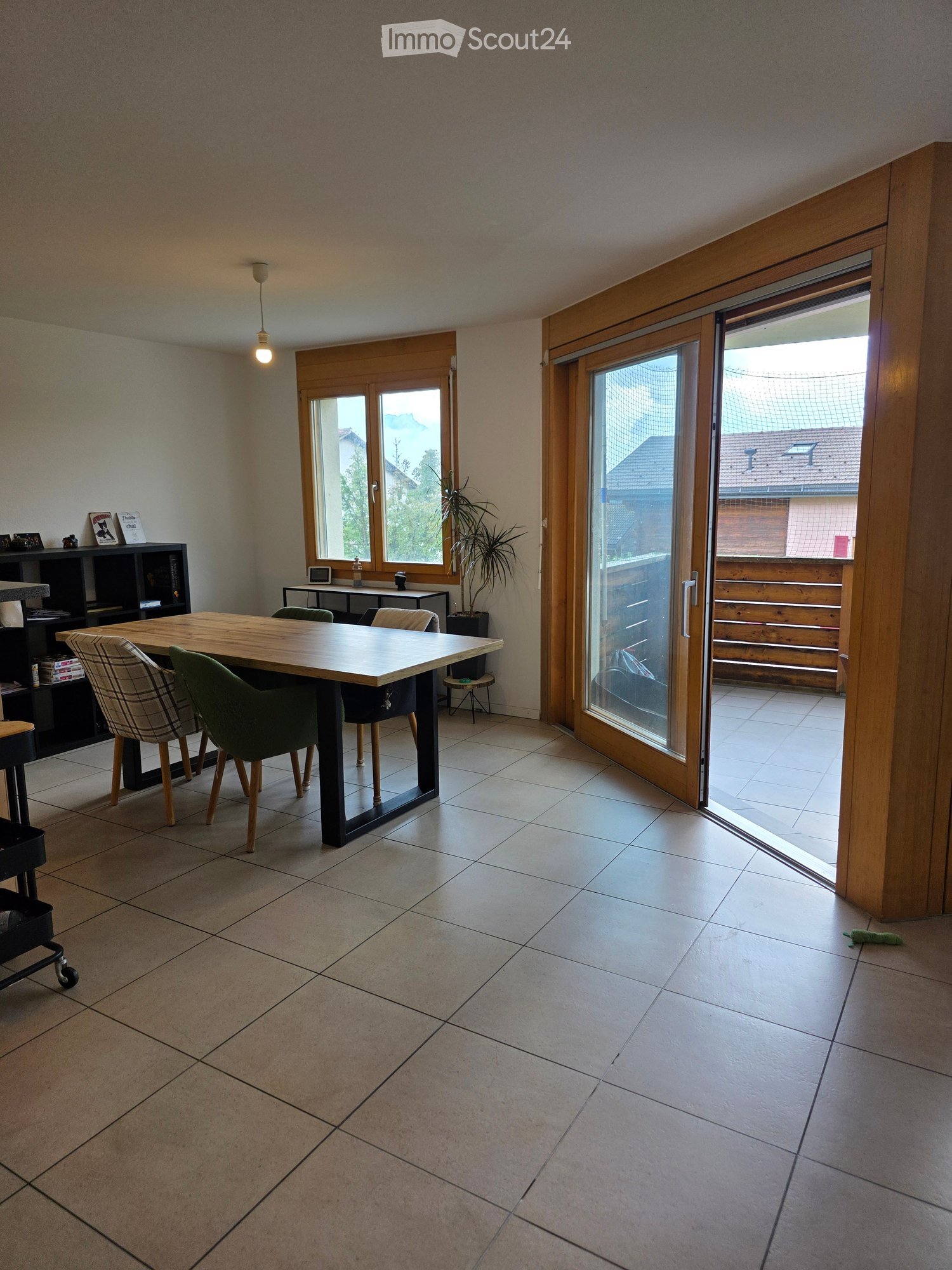 Bright dining area with tiled floor, large wooden dining table, four chairs, a small shelf with decorative items, and a potted plant near the window. A sliding glass door leads to a balcony.