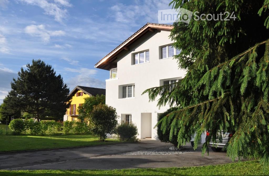 2 story house, white exterior, brown roof, balcony, driveway, car parked, trees, bushes, yellow house in background