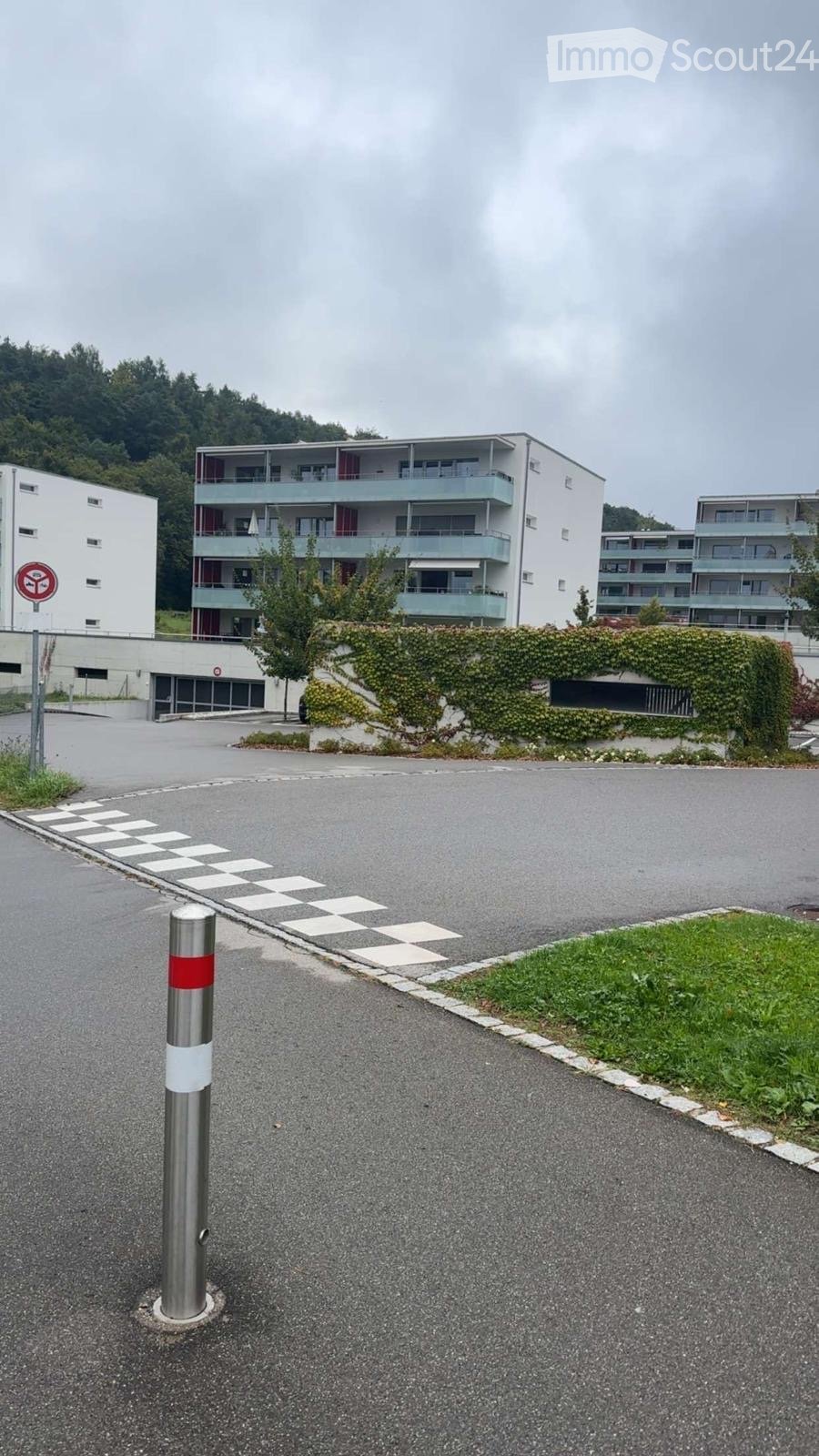 Multi-story residential building with balconies, surrounded by greenery and a paved parking area.