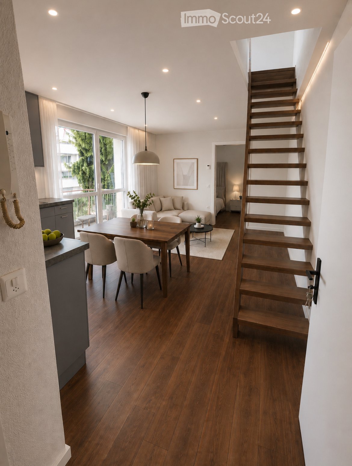 Open-plan living and dining area with wooden flooring, a staircase leading to an upper level, a sofa, dining table with chairs, and a small side table with a lamp.