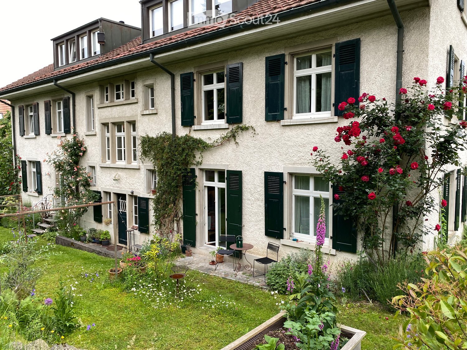 A well-maintained garden with a lawn, various plants, and a small seating area with a table and chairs near the house entrance.