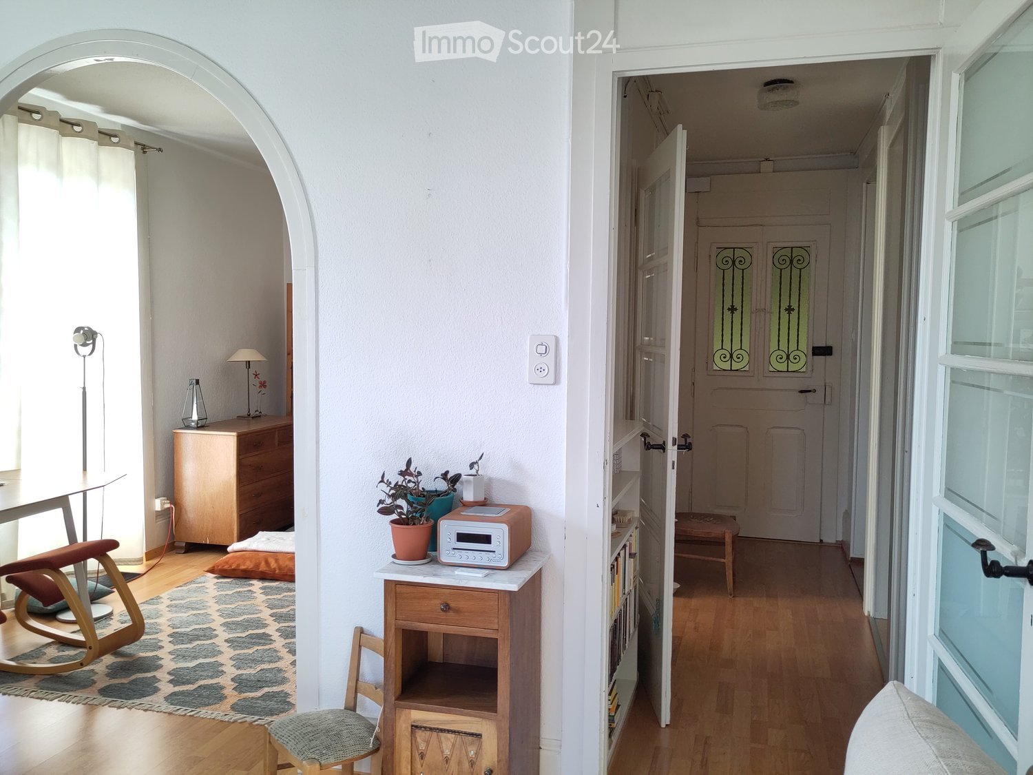 A well-lit hallway with wooden flooring, featuring a small table with a lamp, a cabinet, a rocking chair, and a potted plant on a side table. The hallway leads to another room with a door and radiator.
