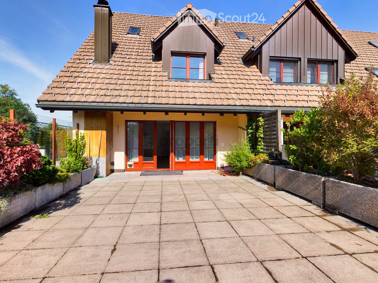 Two-story house with a tiled roof, yellow walls, and red-framed windows. The entrance features large glass doors with curtains, flanked by greenery and a paved walkway.