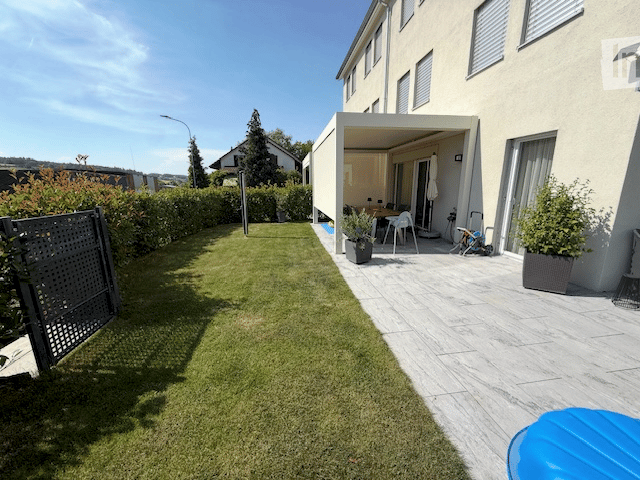 Well-maintained garden with a neatly trimmed lawn, a covered patio area with outdoor furniture, and potted plants. The patio features a table and chairs, and the surrounding area includes a modern house with large windows.