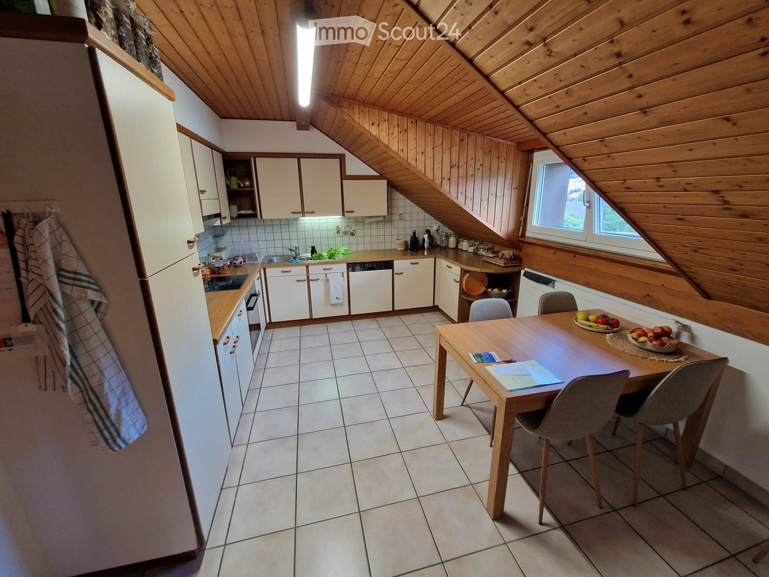 Bright kitchen with white cabinets, wooden ceiling beams, tiled floor, and a dining area with a wooden table and chairs. The kitchen features a refrigerator, dishwasher, and various small appliances on the counter.