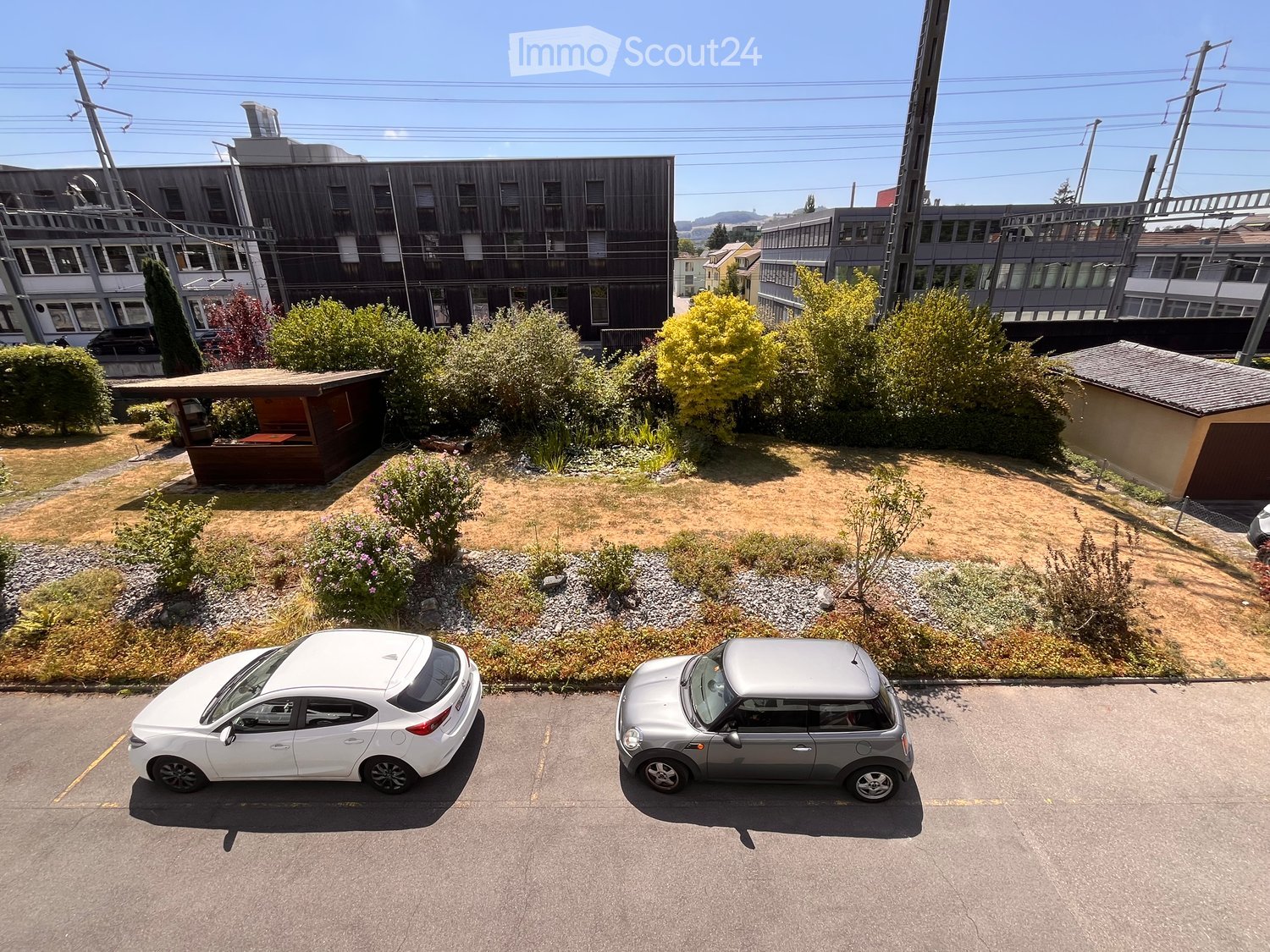 A small garden area with a wooden gazebo, surrounded by bushes and trees, with two parked cars in the foreground.