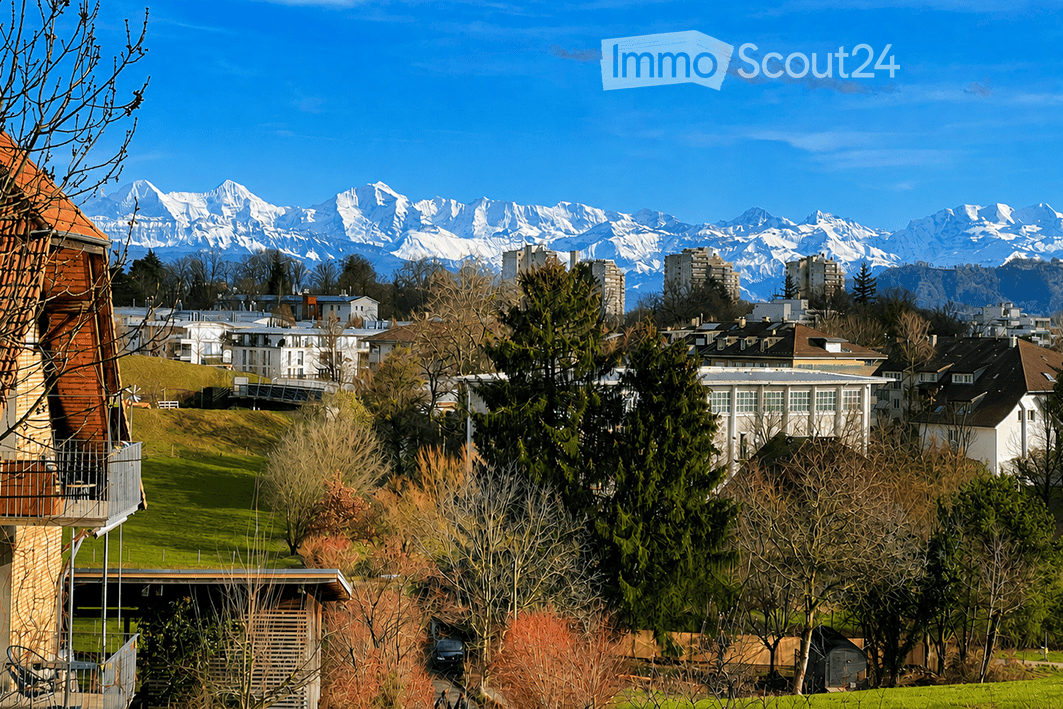 Panoramic view of a city with snow-capped mountains in the background, featuring a mix of residential and commercial buildings, green spaces, and trees.