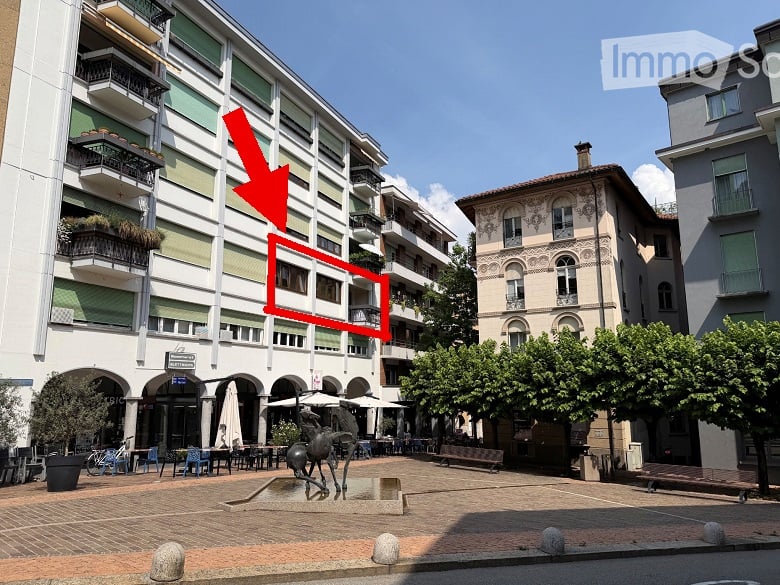 Multi-story apartment building with balconies and green plants, located in a town square with outdoor seating, tables, and a sculpture in the foreground.