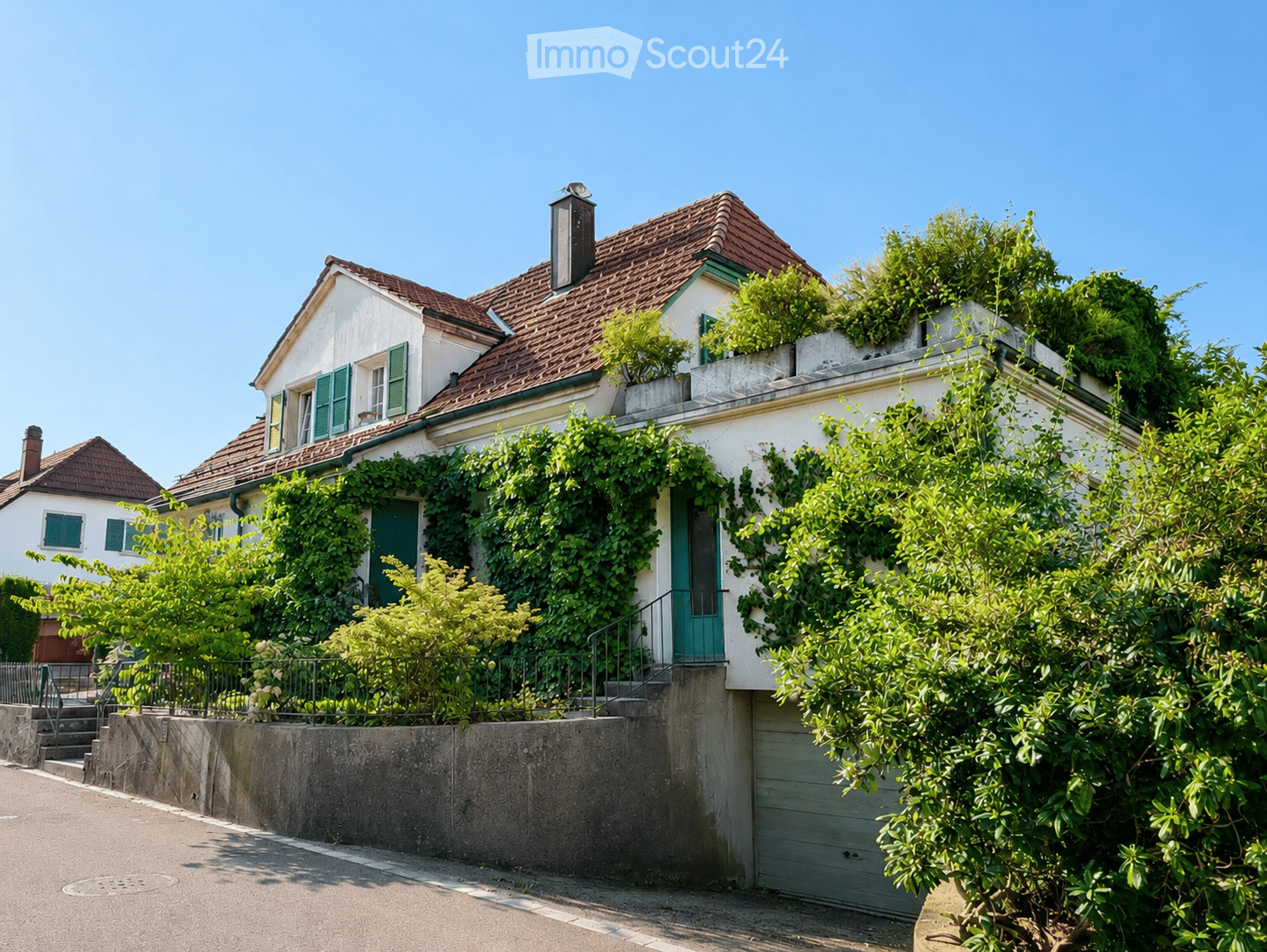 Two-story house with red-tiled roof, white walls, green shutters, and ivy-covered facade. Features a small balcony and a garage entrance.
