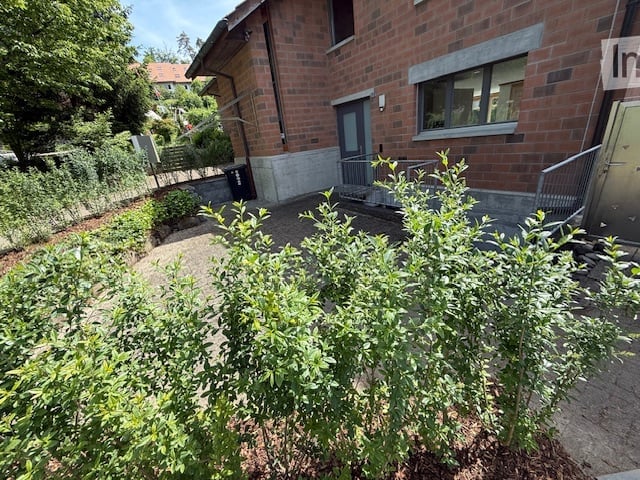 A small garden area in front of a brick building, featuring a gravel pathway, green shrubs, and a small bench. The building has a modern design with large windows and a raised entrance.