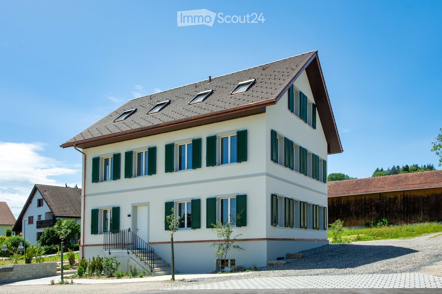 Two-story house with white exterior, green shutters, multiple windows, and a sloped roof with skylights. The house has a raised ground floor with a small staircase leading to the entrance.