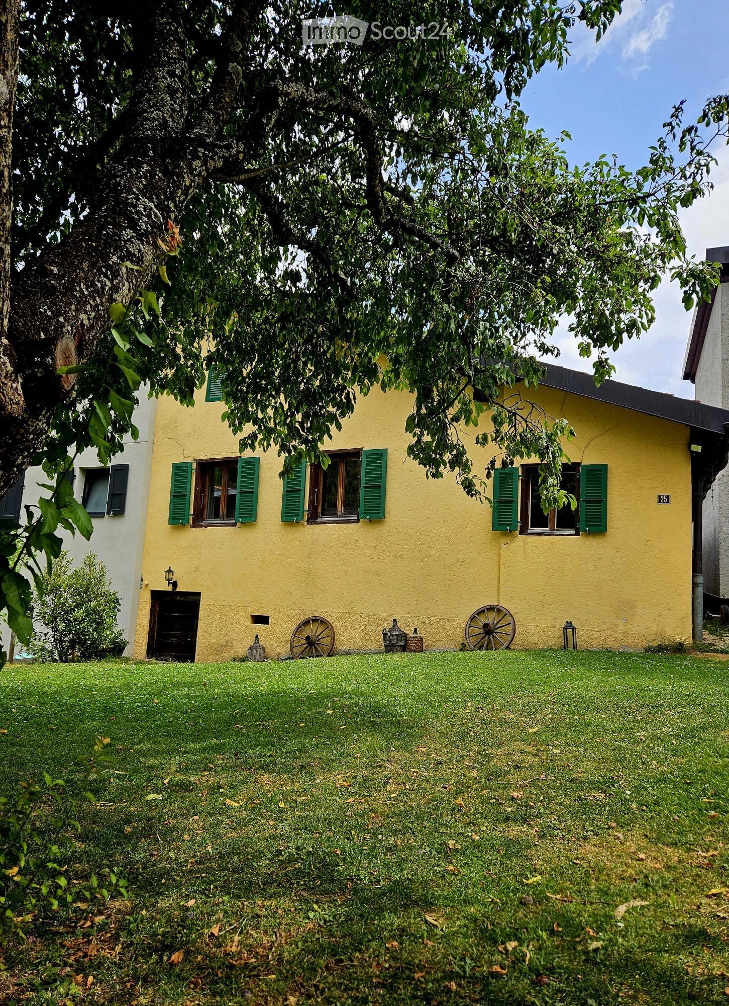 Yellow two-story house with green shutters, a well-maintained lawn, and decorative items like wheelbarrows and baskets near the entrance.