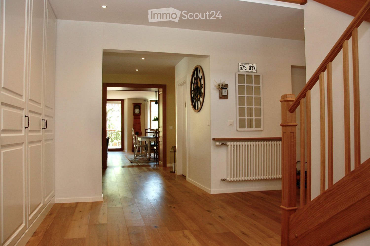 Spacious hallway with wooden flooring, white walls, and a staircase leading upwards. Features a radiator, a wall-mounted clock, and a framed picture. A dining area is visible in the background with a table and chairs.