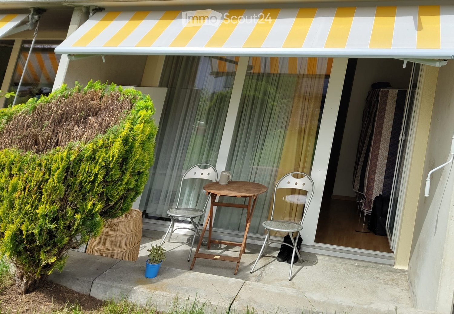 Balcony with a small round wooden table, two white metal chairs, a cup, and a small potted plant. A large sliding glass door leads to the interior, and a yellow and white striped awning provides shade.