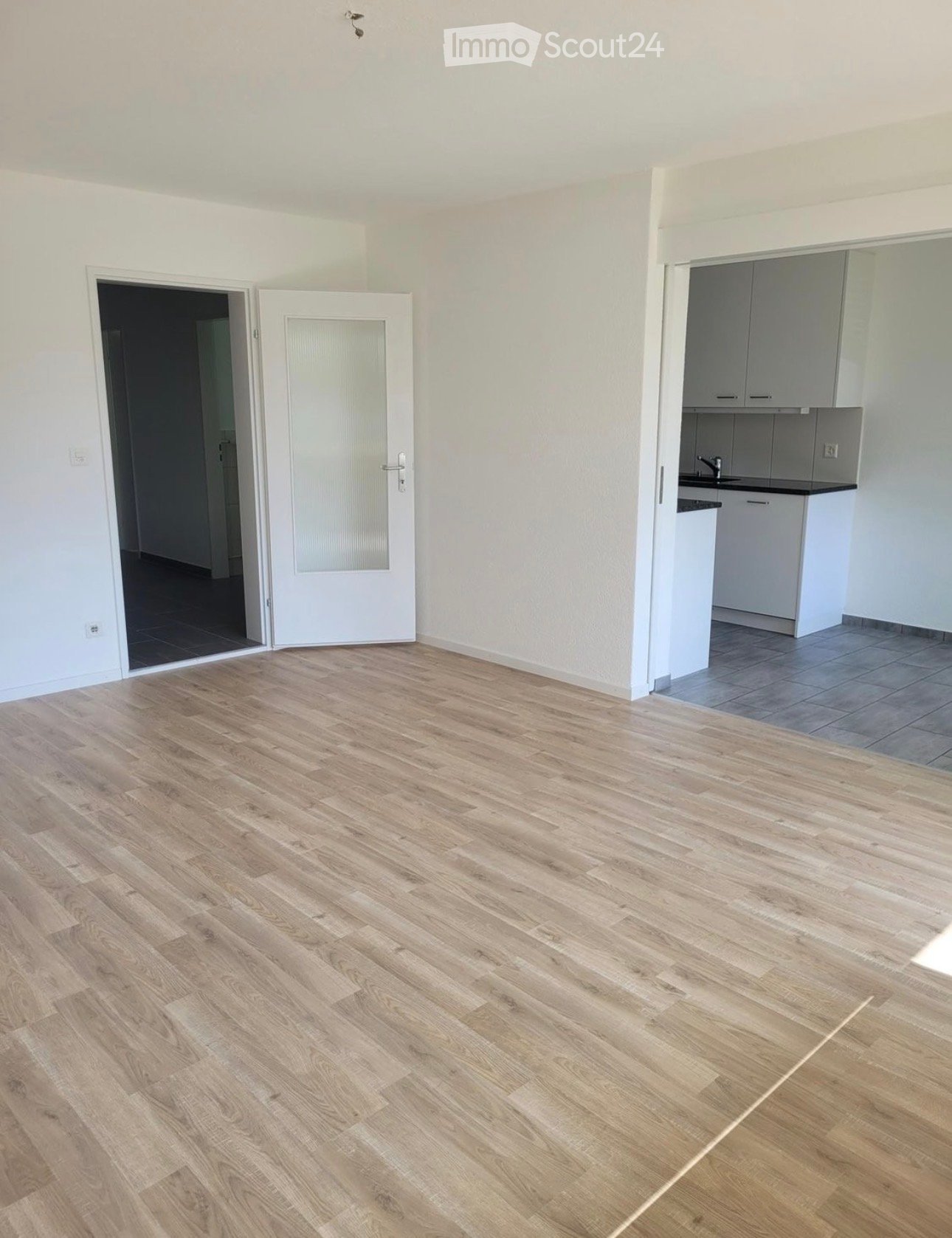 Empty room with light wooden flooring, white walls, and a partially visible kitchen area with cabinets and a sink.