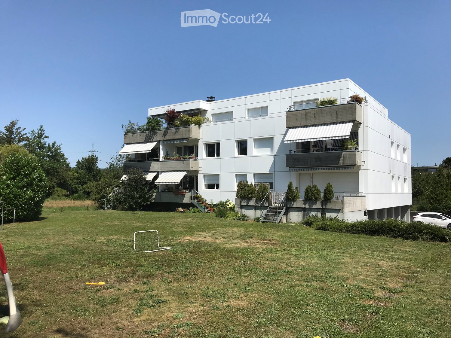 Modern white multi-story building with balconies and greenery, surrounded by a grassy lawn and trees.