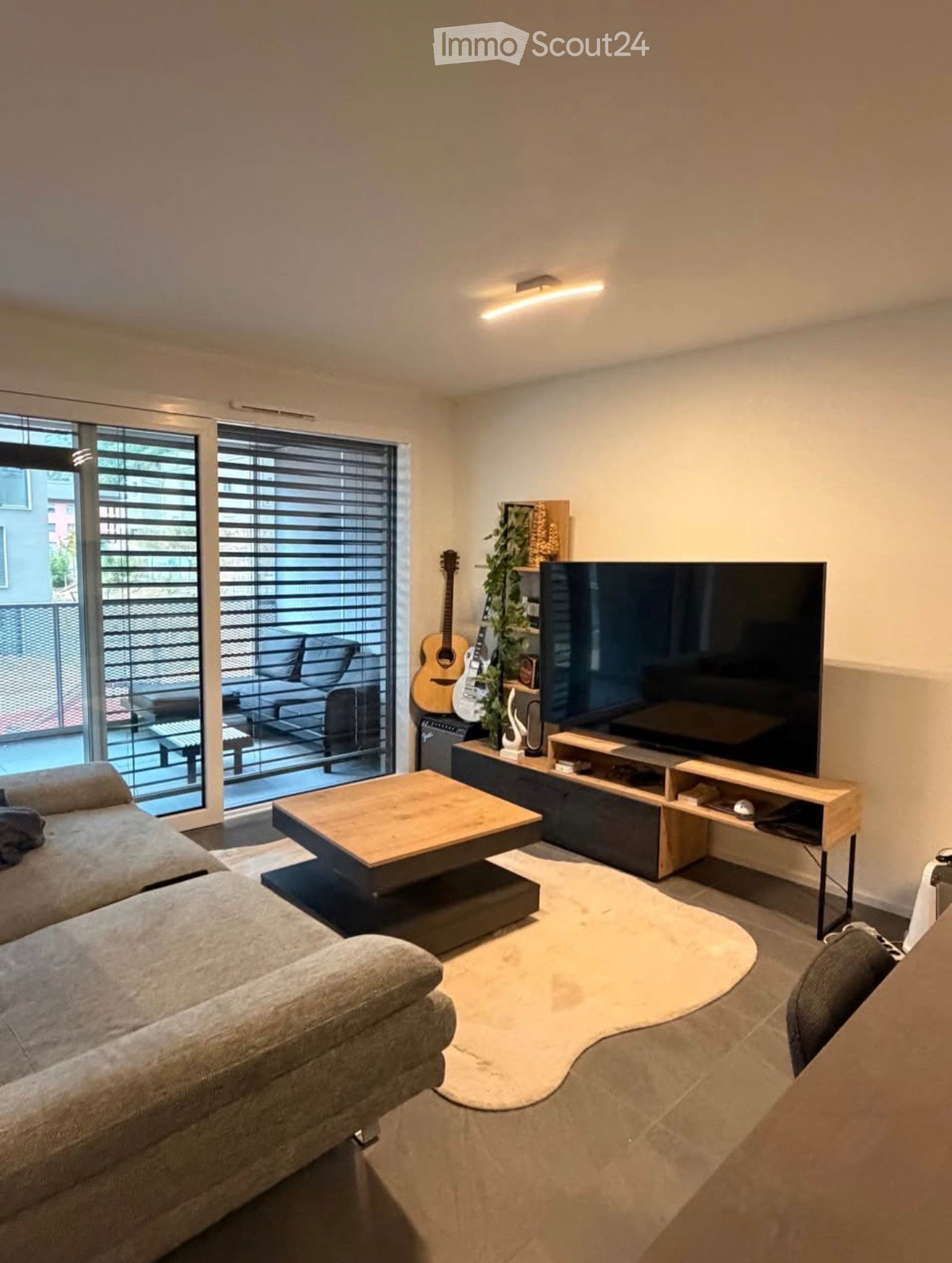 Modern living room with a grey sofa, wooden coffee table, large flat-screen TV, and a guitar leaning against the wall. The room features large windows with blinds and a view of a balcony area.