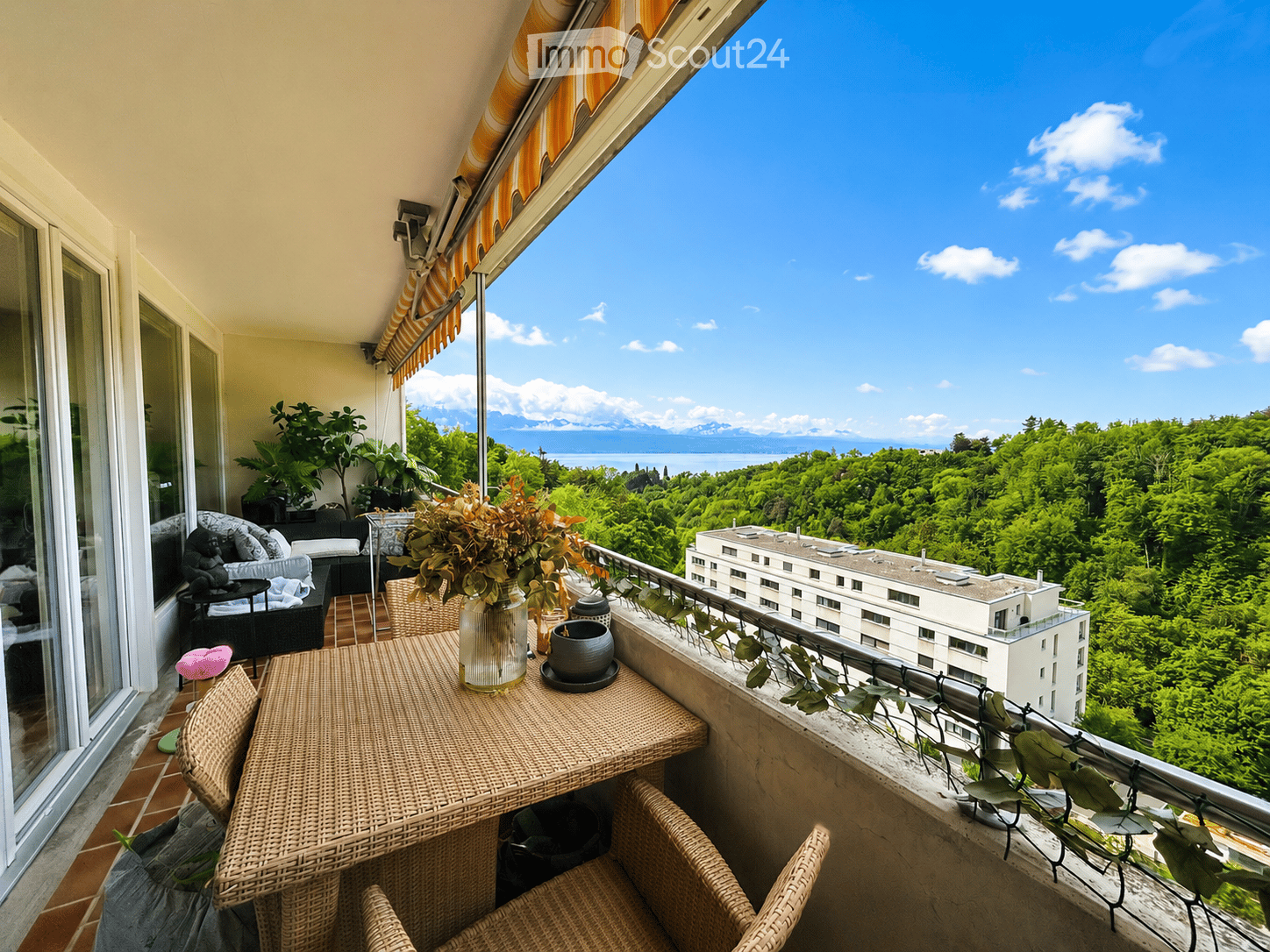 Balcony with wicker furniture, vase with flowers, scenic view of lake and mountains.