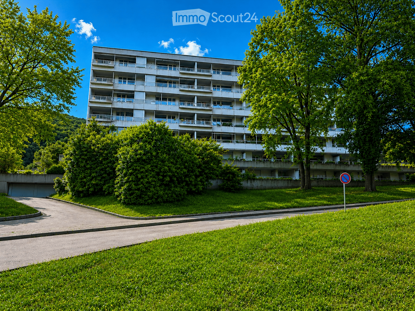 Multi-story residential building with balconies, surrounded by greenery and a paved pathway.