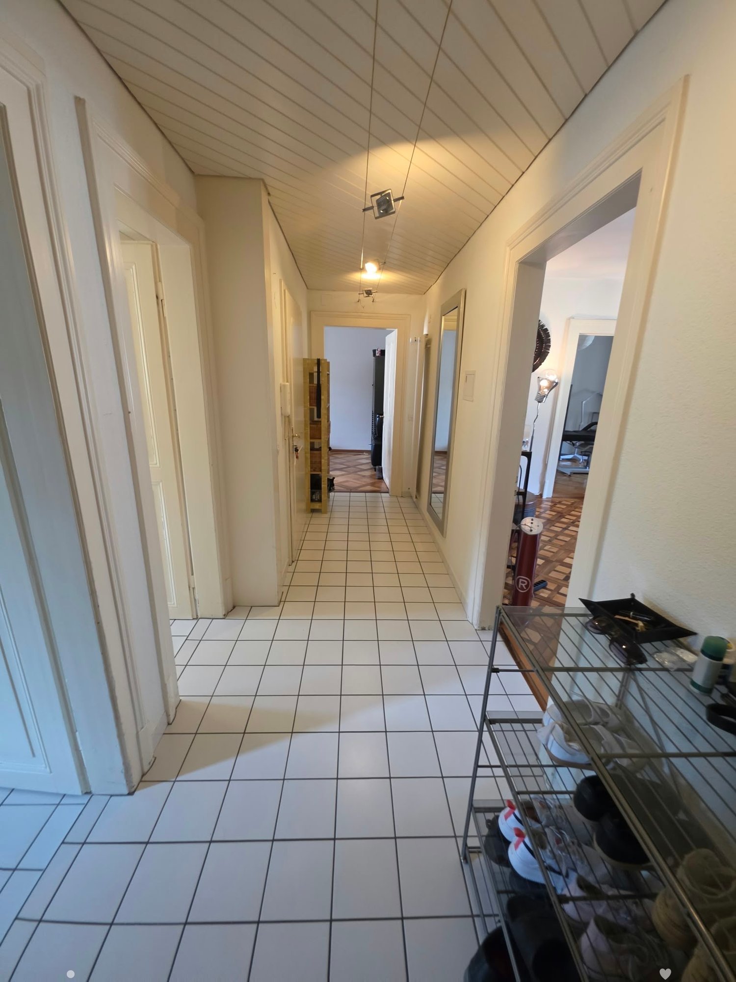 Clean hallway with white tiled flooring, white walls, and multiple doors leading to different rooms. A small metal shelf with shoes and items is visible on the right side.