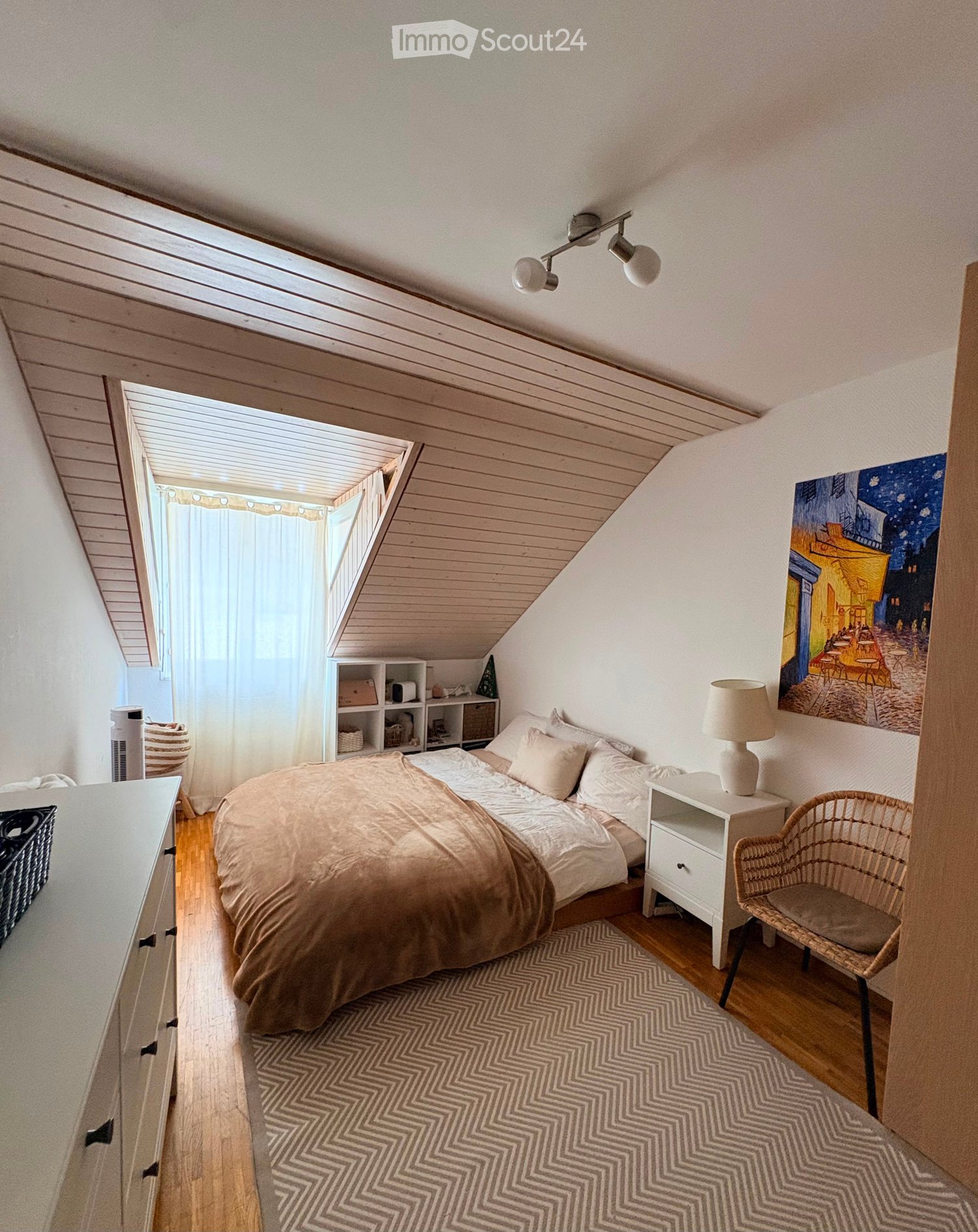 Cozy attic bedroom with wooden slanted ceilings, large skylight, bed with beige bedding, nightstand with lamp, wicker chair, shelf, and artwork on the wall.