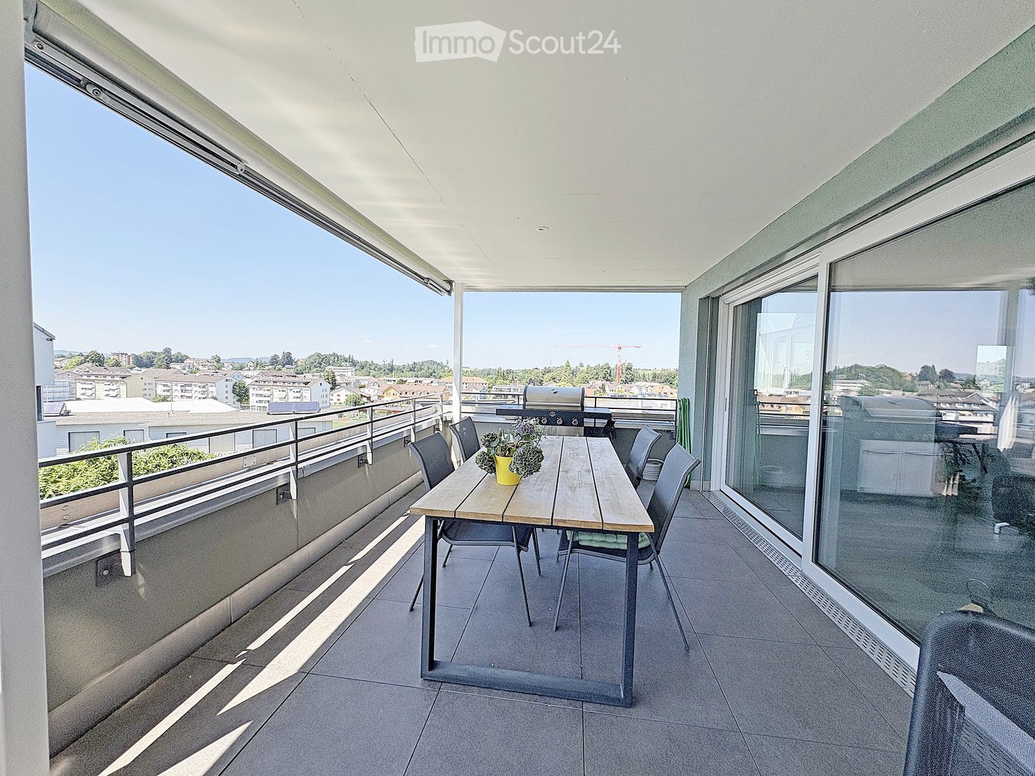 Balcony with a wooden table, chairs, and a potted plant, offering a view of the cityscape.