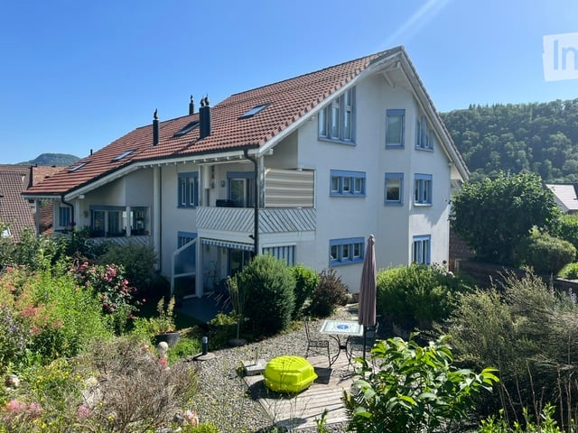 Two-story house with white exterior, red-tiled roof, multiple windows, and a small balcony. The surrounding garden features a patio with outdoor furniture and plants.