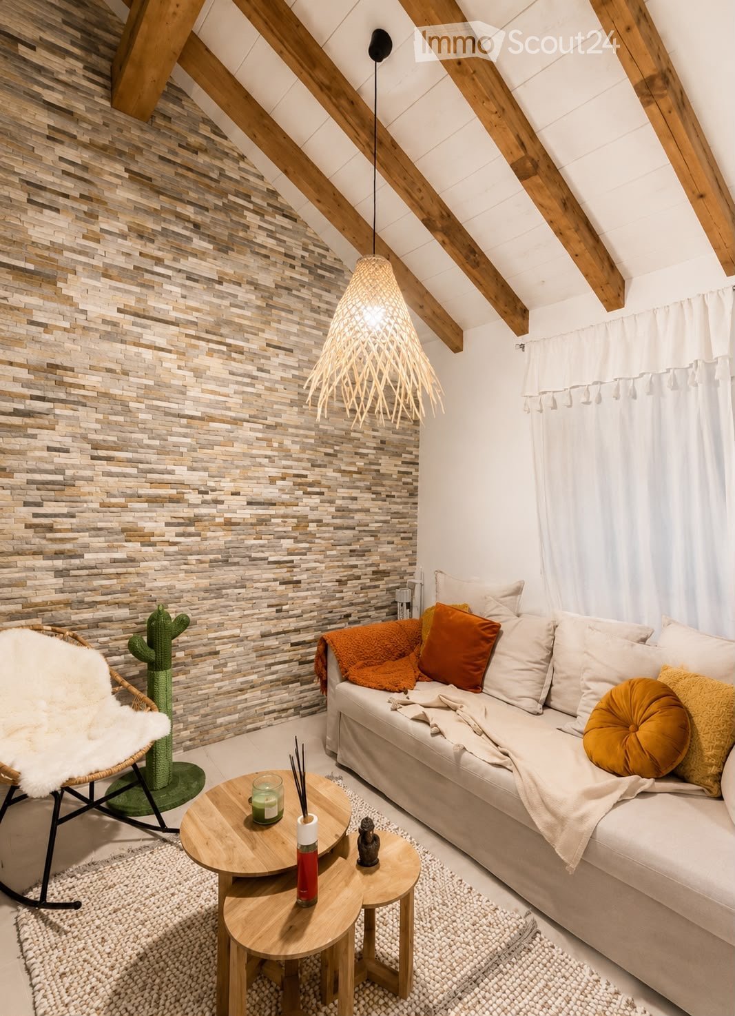 A cozy living room with exposed wooden beams, a textured stone accent wall, a white sofa with orange and yellow cushions, a wooden coffee table, a wicker chair with a white fur throw, and a hanging woven pendant light.