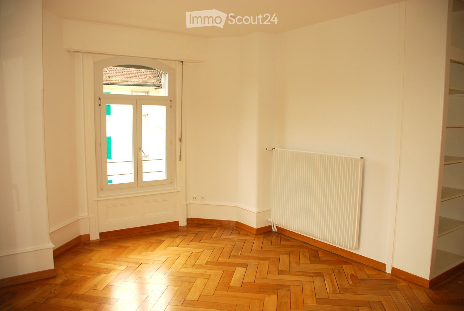 Empty room with wooden herringbone flooring, white walls, a radiator, and a window with a view of a roof.
