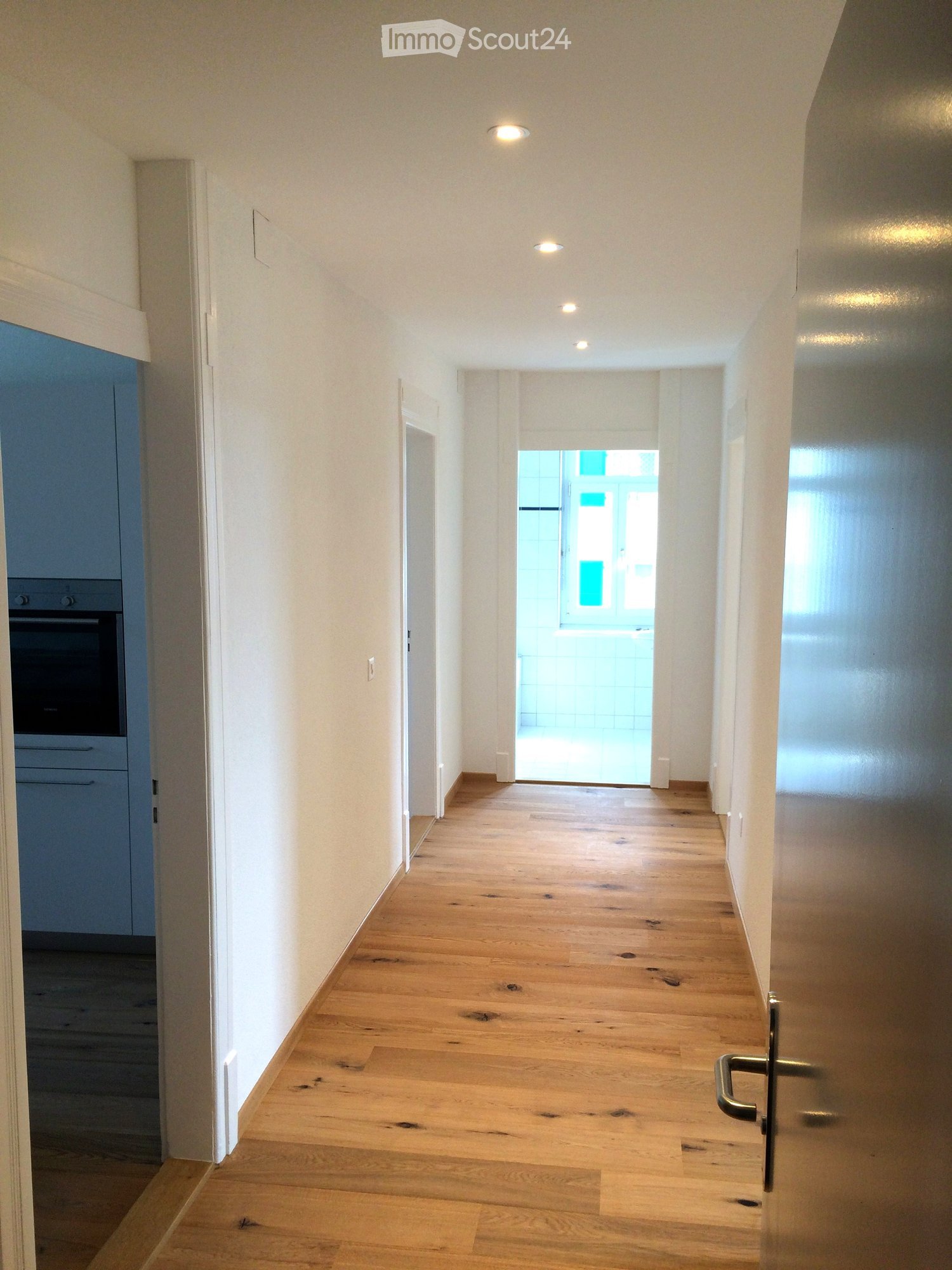 Empty hallway with light wooden flooring, white walls, and recessed ceiling lights. The hallway leads to an open area with a view of a tiled space beyond.