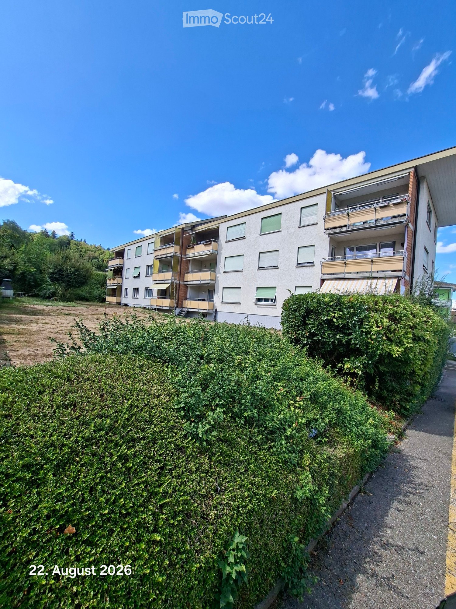 Multi-story residential building with balconies, surrounded by greenery and a pathway.