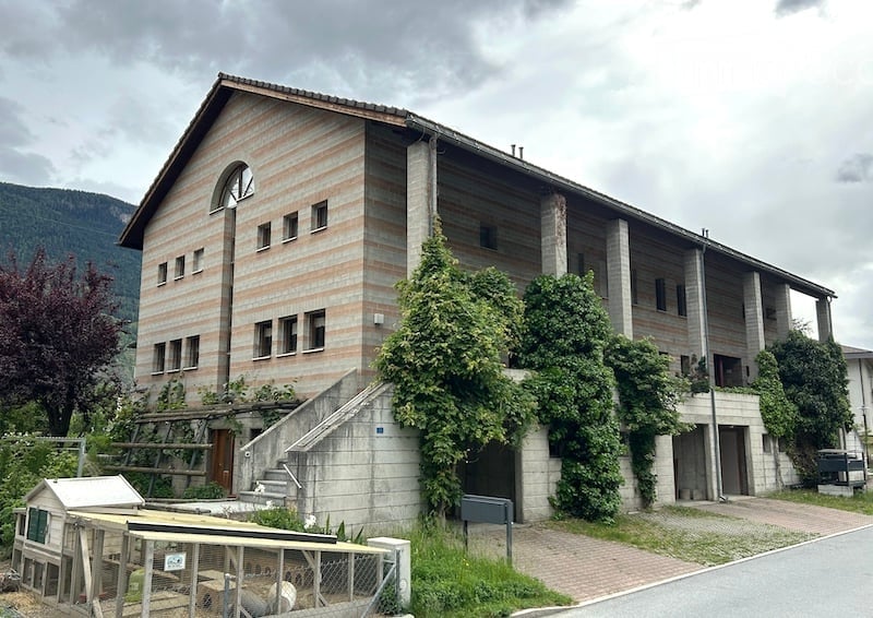 Two-story building with a striped facade, multiple windows, and a staircase leading to the entrance. The building is surrounded by greenery and a small fenced area.