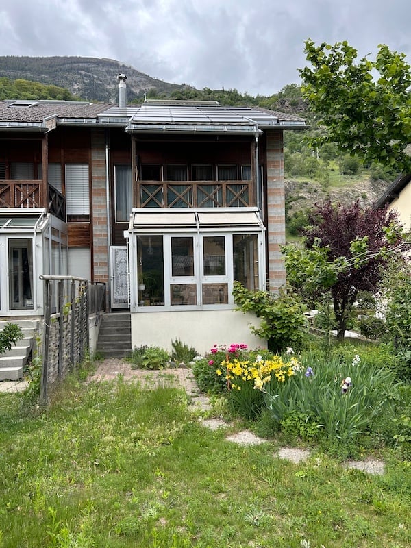 Two-story house with a balcony, surrounded by a garden with colorful flowers and a mountain view in the background.