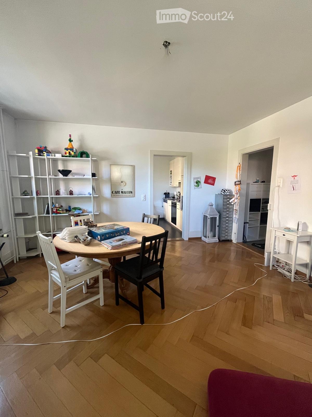 A well-lit dining area with a round wooden table, chairs, a white shelf with decorative items, and a view into a small kitchen with white cabinets, a refrigerator, and an oven.