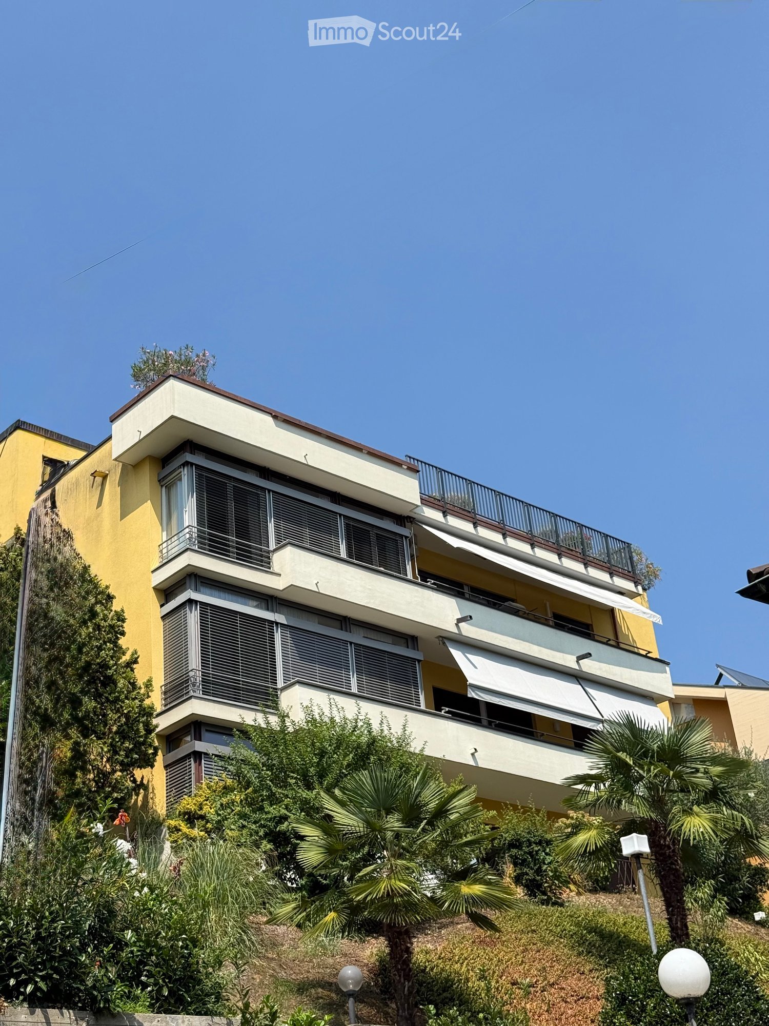 Multi-story residential building with balconies, large windows, and surrounding greenery including palm trees and shrubs.