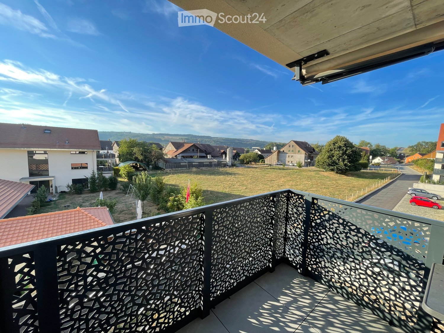 Balcony with modern black metal railing, overlooking a residential area with houses, trees, and a clear blue sky.